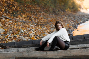 A girl is sitting on a wooden boat. In the background you can see a river bank covered with autumn leaves and stones. The water in the river reflects golden light.