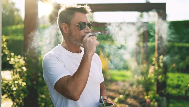 Middle-aged man smoking cigar in garden. Senior businessman relaxing, celebrating the end of successful business day. Mature male in sunglasses enjoying sunset lights on terrace of country home. - Powered by Adobe
