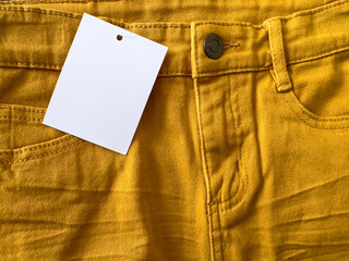 New mustard pants with white label, new item. Blank price tag on jeans. Label on clothes. New orange jacket with white price tag. Fragment of orange jeans. Sewing clothes