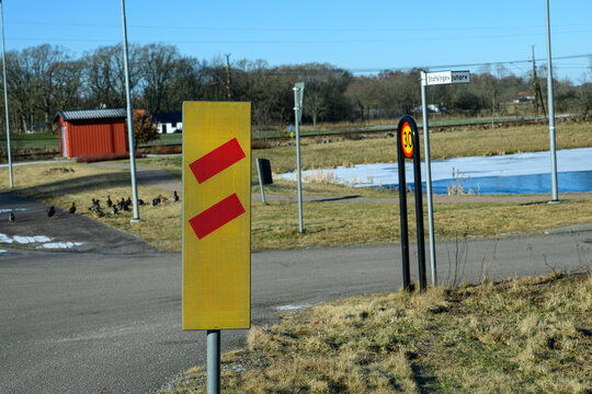 A bright yellow road sign with red chevrons alerts drivers closing railroad. In the background, a red building and ducks can be seen in a peaceful rural setting