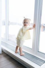 Vertical shot of adorable toddler girl with two playful ponytails standing by window on windowsill, soaking in winter scenery enjoying cozy warmth of home. Charming scene of childhood innocence.