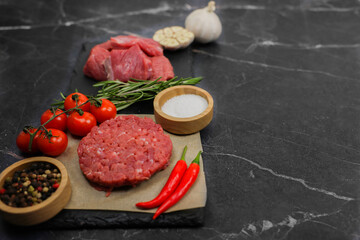 Close-up on the left is a raw beef round patty lying on paper surrounded by seasonings and vegetables, behind them separated by a sprig of rosemary lies meat cut into chunks on a dark background
