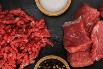 A close-up view from above of chunks of beef meat and red minced meat with streaks of fat lying on a black background with spices and salt in wooden bowls on the sides. The concept of cooking