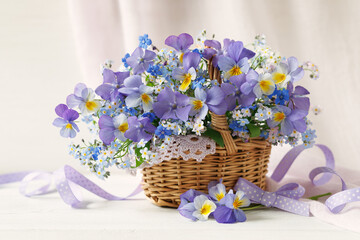 Spring forget-me-not flowers, viola pansy, periwinkle in a basket, bouquet with ribbon on a white table.
