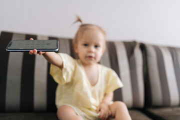 Toddler girl looking at camera sitting on sofa, holding smartphone with cracked screen showing it forward, potentially indicating early exposure to technology or seeking attention, selective focus.