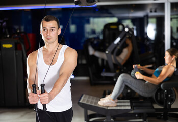 Fototapeta premium Slender athletic man work out muscles of arms and shoulders on TRX rope loop simulator in gym of fitness club. Gym visitor trains triceps during classes at multi-station in wellness center.