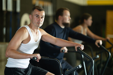 Determined young guy training with air bike in sports hall during crossfit workout