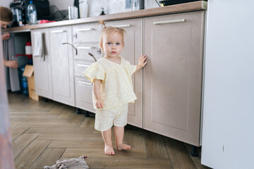 Full length portrait of curious toddler girl in yellow outfit exploring kitchen cabinets, touching...