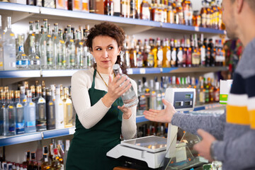 Adult saleswoman in uniform sells bottle of vodka to adult male customer in grocery store