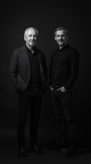 Fototapeta premium Two men, one older and one younger, stand side-by-side against a dark background. A monochrome portrait.