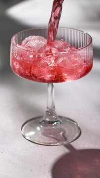 fresh red drink pouring into a cocktail glass with ice cubes on a gray sunny restaurant tabletop, slow motion