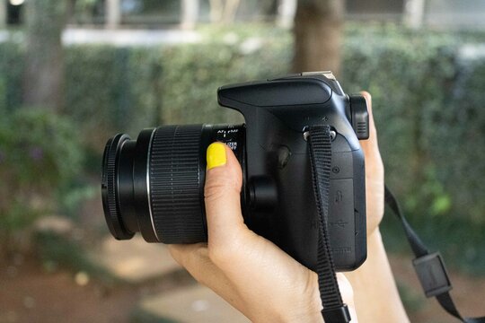 M&atilde;o com unhas pintadas com esmalte de cor amarela segurando uma c&acirc;mera fotogr&aacute;fica DSLR. 