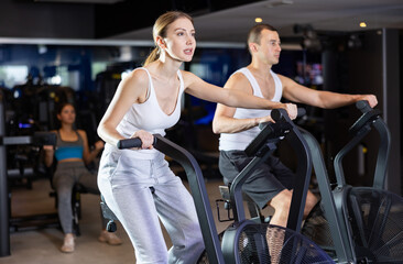 Sportive guy and young girl engaged in training with airbike machine in exercise room