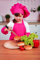 Funny happy little beautiful girl in a chef costume prepares burgers while sitting at a table in the kitchen. Healthy eating.