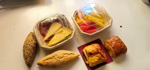Assorted breakfast items on a white table; two plastic containers with sliced cheese, vegetables, and fruit; three types of bread; two pastries on a red napkin;