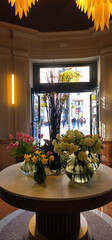 A round table with glass vases filled with tulips, daffodils, and white hydrangeas; an open doorway in the background shows people walking on the street;