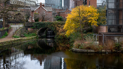 A peaceful view of a historic canal in Manchester, surrounded by autumn foliage and charming red brick buildings. A small waterfall flows beneath a stone bridge covered