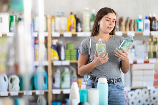 Young female customer in home essentials store holding two bottles of dishwashing liquid, carefully comparing product features before making decision