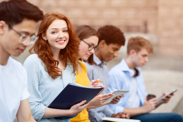 Learning Concept. Portrait of cute red-haired girl studying outdoors with her friends and looking at camera