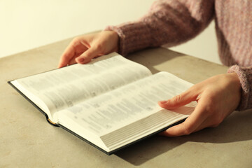 Woman reading Holy Bible in English language at beige table, closeup