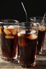Refreshing cola with ice cubes and drinking straws in glasses on wooden table against black background, closeup