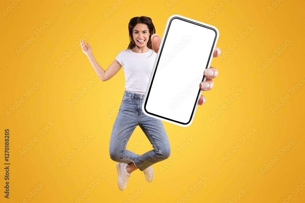 Poster overjoyed young casual lady showing empty smart phone screen while jumping up over yellow studio bac