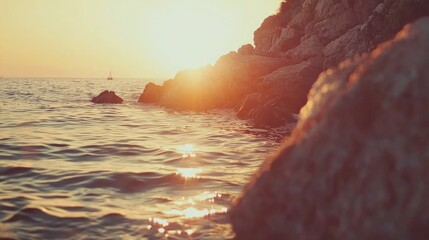 Serene sunset over a tranquil sea with rocks in the foreground and a distant sailboat