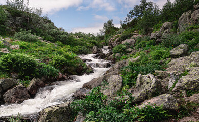 A serene stream cascading over rocky terrain in a vibrant Nordic landscape. The lush greenery and blue cloud sky create a peaceful and inviting atmosphere in nature
