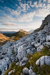 Fototapeten Rocky mountain landscape with scattered clouds in the sky and a golden hued peak in the distance © Boragoo