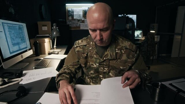 Medium shot of Caucasian man in military uniform signing documents while sitting at desk with computer in intelligence office