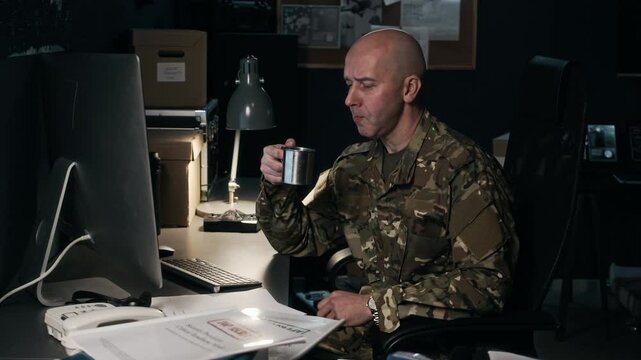 Medium shot of adult Caucasian military analyst doing report on computer while sitting at desk with folders and documents and drinking tea