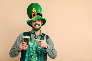 Young man in leprechaun hat with green beard pointing at glass of beer on beige background. St....