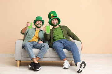 Happy young men in leprechaun hats with green beards holding glasses of beer and sitting on sofa...