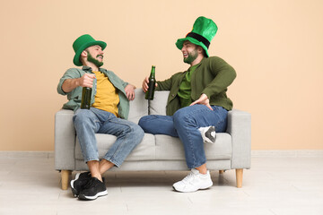 Young men in leprechaun hats with green beards holding bottles of beer and sitting on sofa near...