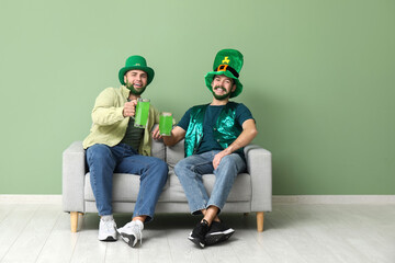 Young men in leprechaun hats with green beards holding glasses of beer and sitting on sofa near...