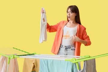Shocked young woman with stained clothes and dryer on yellow background
