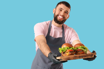 Young male chef holding wooden tray with tasty burgers on blue background