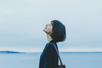 Fototapeta premium A Moment of Serenity: A young woman gazes skyward with a smile, embracing the gentle breeze beside a calm sea, lost in a moment of peace and tranquility.
