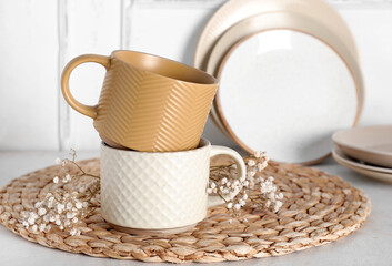Different ceramic mugs with dried flowers and wicker mat on light background