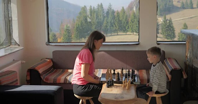 Young woman and her little son play chess in a mountain rustic chalet