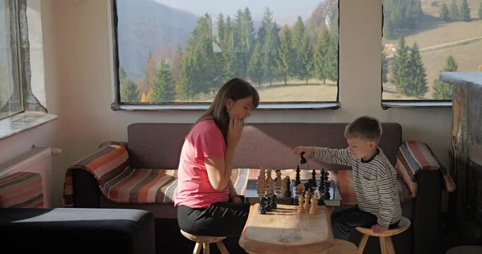 Little kid and his mother play chess in a mountain rustic chalet
