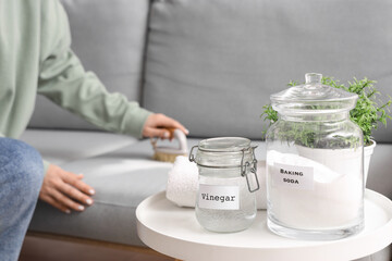 Jars of baking soda and vinegar on table against woman cleaning sofa at home, closeup