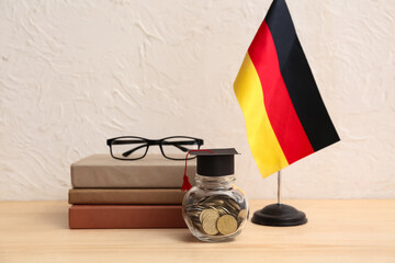 Jar of money with graduation hat, books and German flag on table near light wall. Student loan...