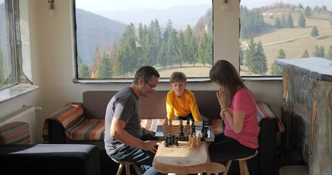 Young couple playing chess in a mountain rustic chalet, kid arbitrates
