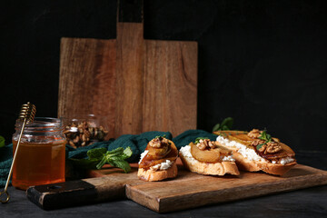 Wooden board of pear bruschetta with ricotta cheese, walnuts and jar of honey on black background
