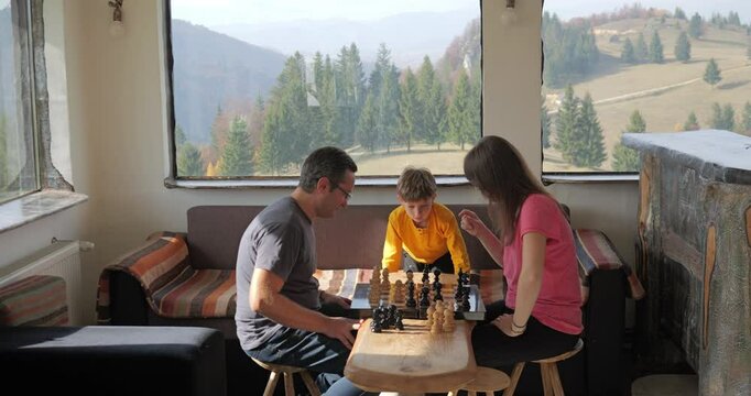 Man and woman playing chess in a mountain rustic chalet, child arbitrates