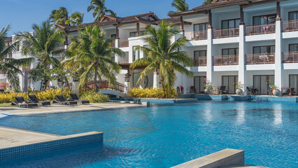 Swimming pool at the resort. Ripples on a blue water. Sun beds in the recreation area. A lifebuoy on the railing of an arched pedestrian bridge. The hotel building has balconies, terraces. Philippines