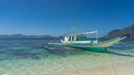 Fototapeta premium A beautiful traditional double-outrigger dugout Filipino bangka boat is moored at the shore. The ropes are stretched. The turquoise ocean water is crystal clear. Mountains on the horizon. Blue sky. 