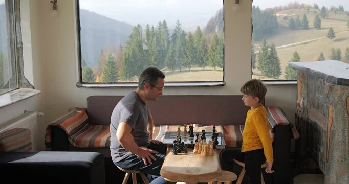 Father and son play chess in a rustic mountain cottage, checkmate game over boy winner shake hand with man