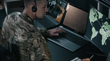 Back view of young male military officer in uniform wearing headset transmitting data while using laptop and computer with screen of global map